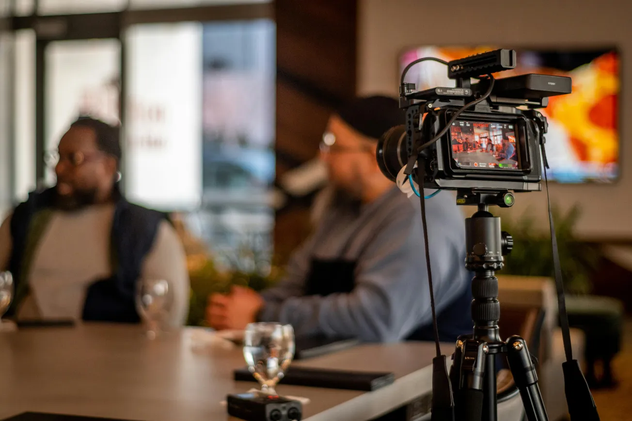 Camera on a tripod focuses on two people sitting at a table for an interview. Blurred background shows a casual setting with ambient lighting.