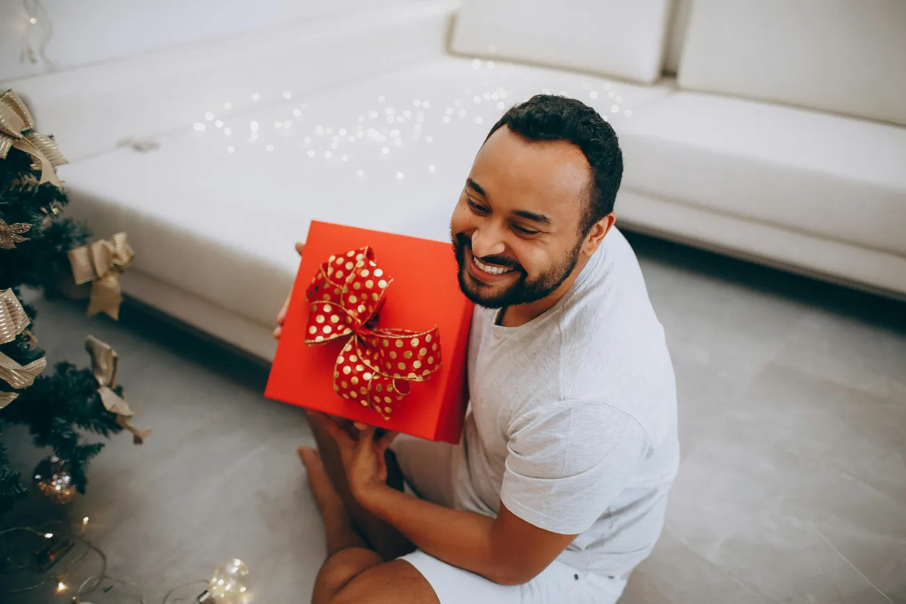A man smiles while holding a bright red gift with a large, red bow with gold polka dots. A decorated Christmas tree and lights adorn the cozy room.