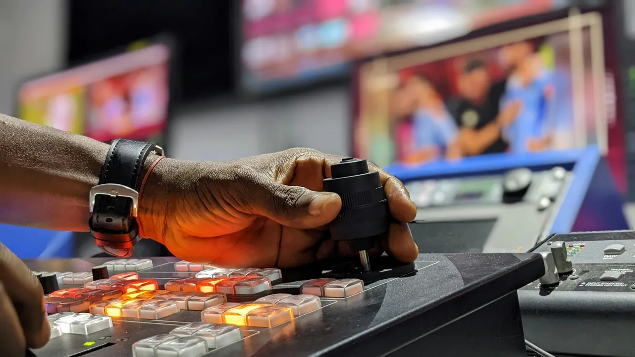 A hand operates a video control board with glowing buttons, set against blurred monitors displaying sports footage, conveying a focused, technical atmosphere.