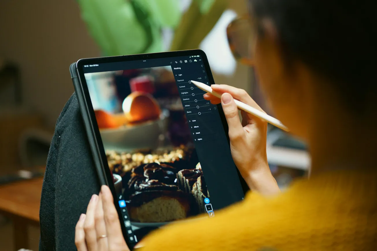 A person uses a stylus on a tablet to edit a photo of pastries and fruit. Warm, creative atmosphere, focusing on editing tools and gestures.