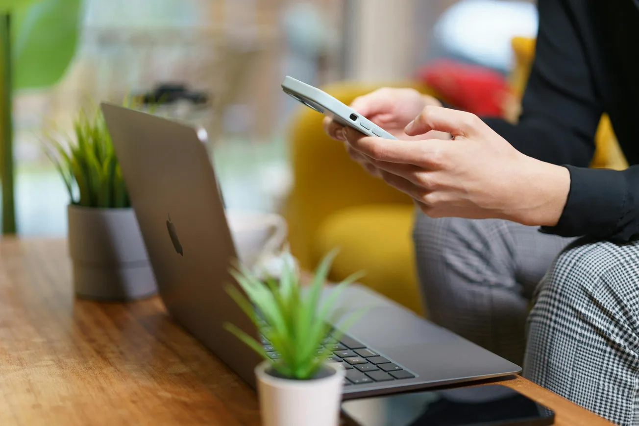 A person sitting on a yellow couch checks their smartphone beside an open laptop on a wooden table. Green plants add a fresh, calm vibe.