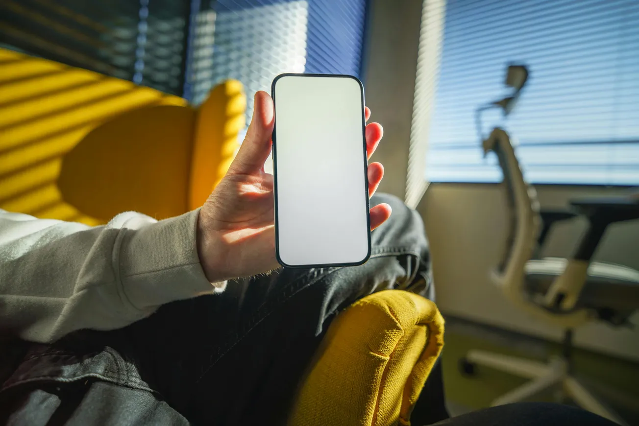 A person holds a smartphone with a blank screen in a sunlit room. They're seated on a yellow chair, with blinds casting striped shadows. A chair is in the background.