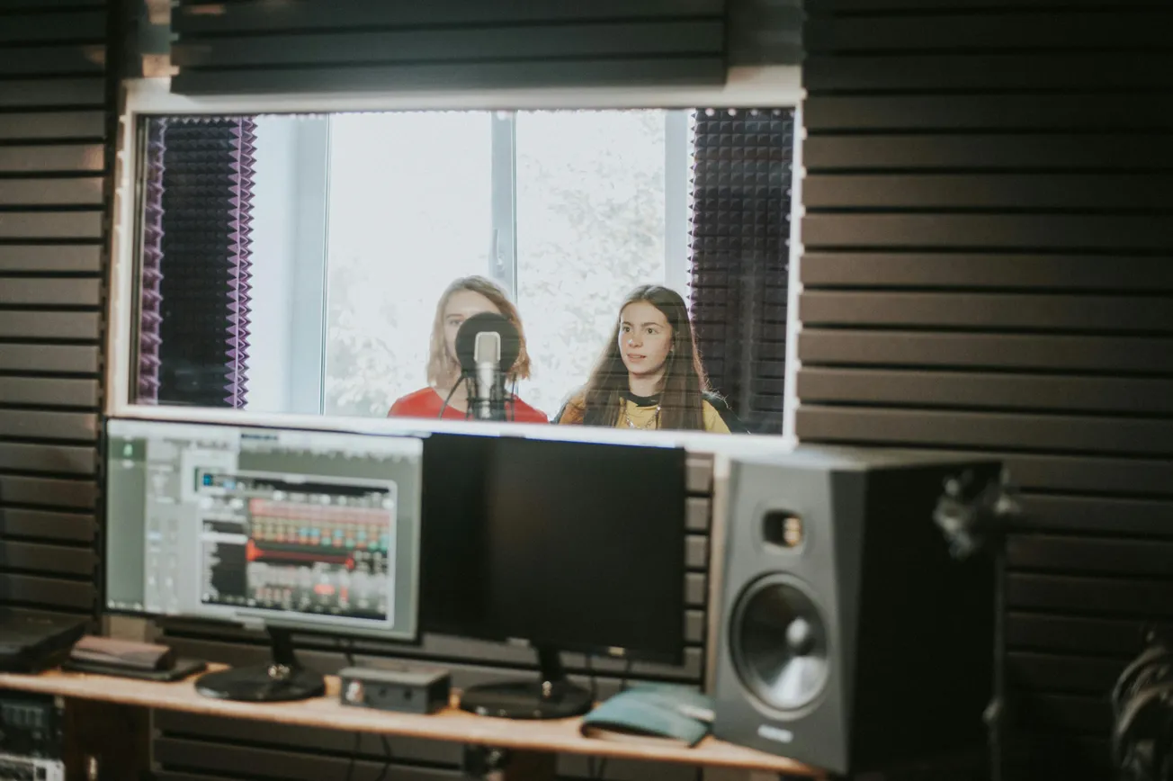 Two people stand behind a microphone in a recording booth, visible through a window. The studio has monitors and crisp acoustics, creating a focused, professional atmosphere.