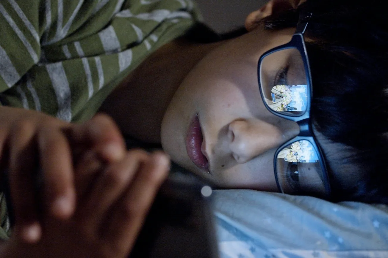 A child in glasses lies in bed, wearing a striped shirt, focused on a smartphone. The phone's screen reflects in their glasses, suggesting nighttime browsing.