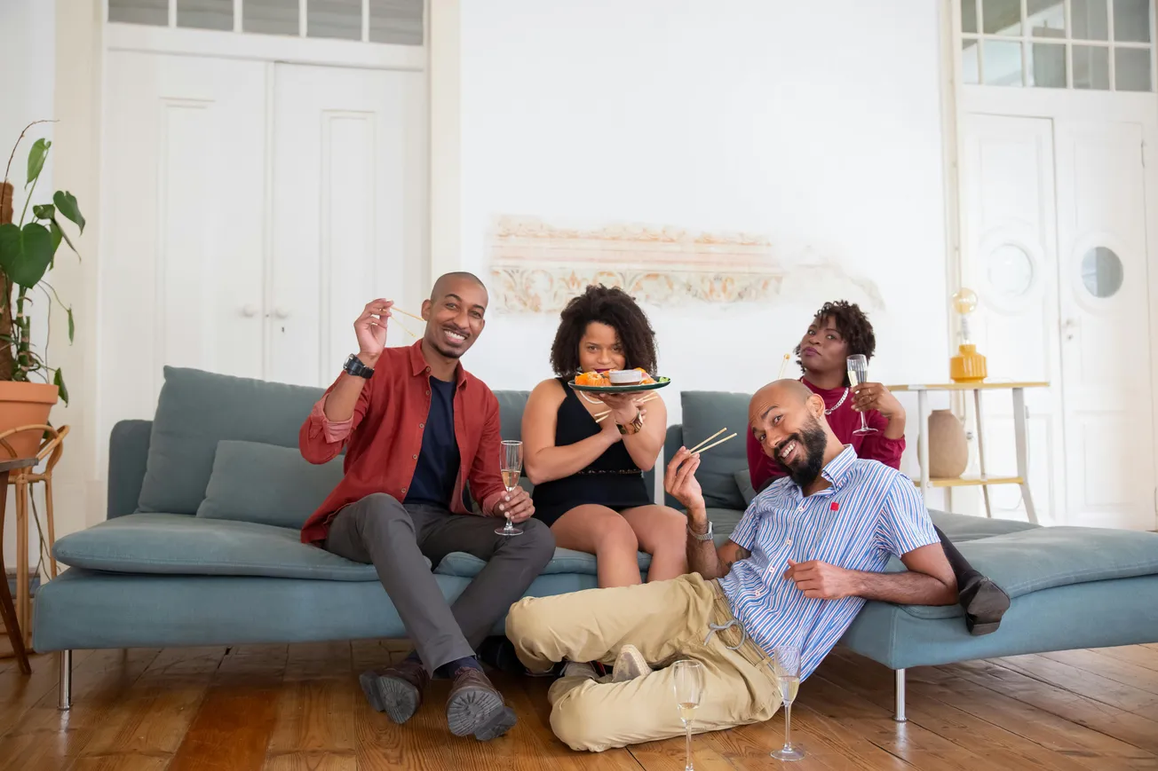 Four people are gathered on a couch, smiling and enjoying sushi. They hold champagne glasses, conveying a cheerful, relaxed atmosphere in a cozy living room.
