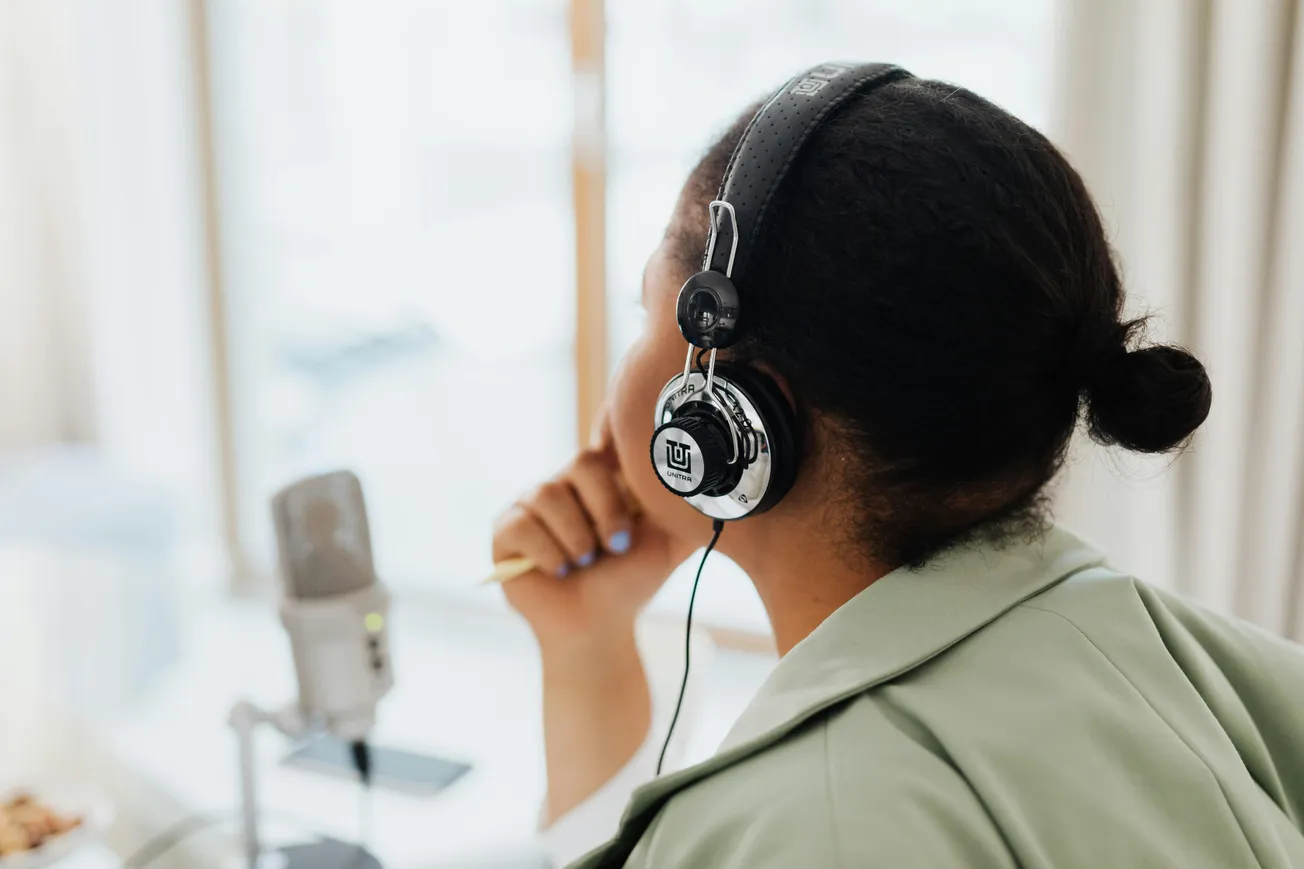A woman wearing headphones sits thoughtfully at a desk with a microphone. Her fingers rest on her chin, suggesting focus and concentration.