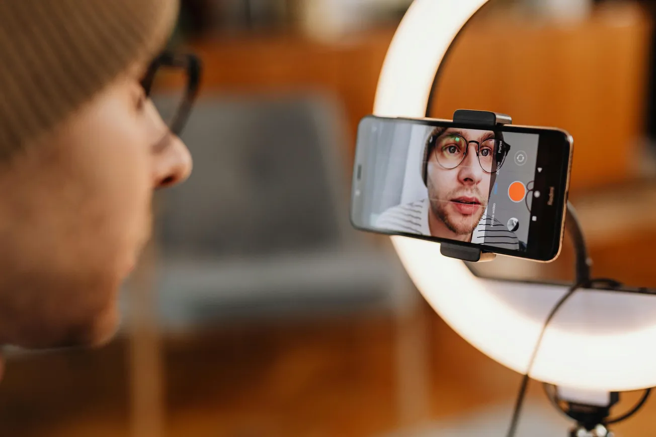 A person wearing glasses and a beanie records themselves with a smartphone mounted on a tripod and ring light, creating a focused, professional setup.