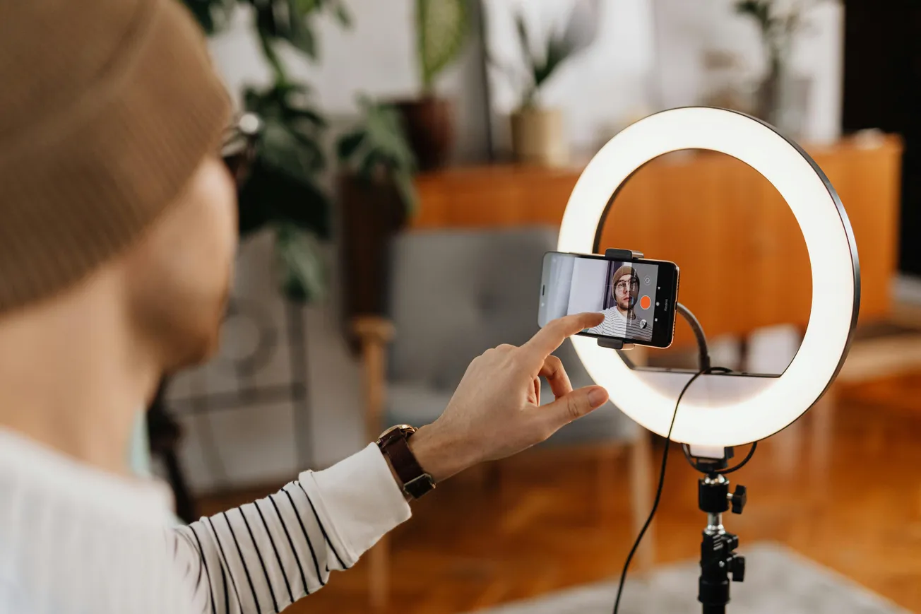 A person wearing a beanie is adjusting a smartphone, mounted inside a ring light on a tripod, likely recording a video. The room is warmly lit with plants.