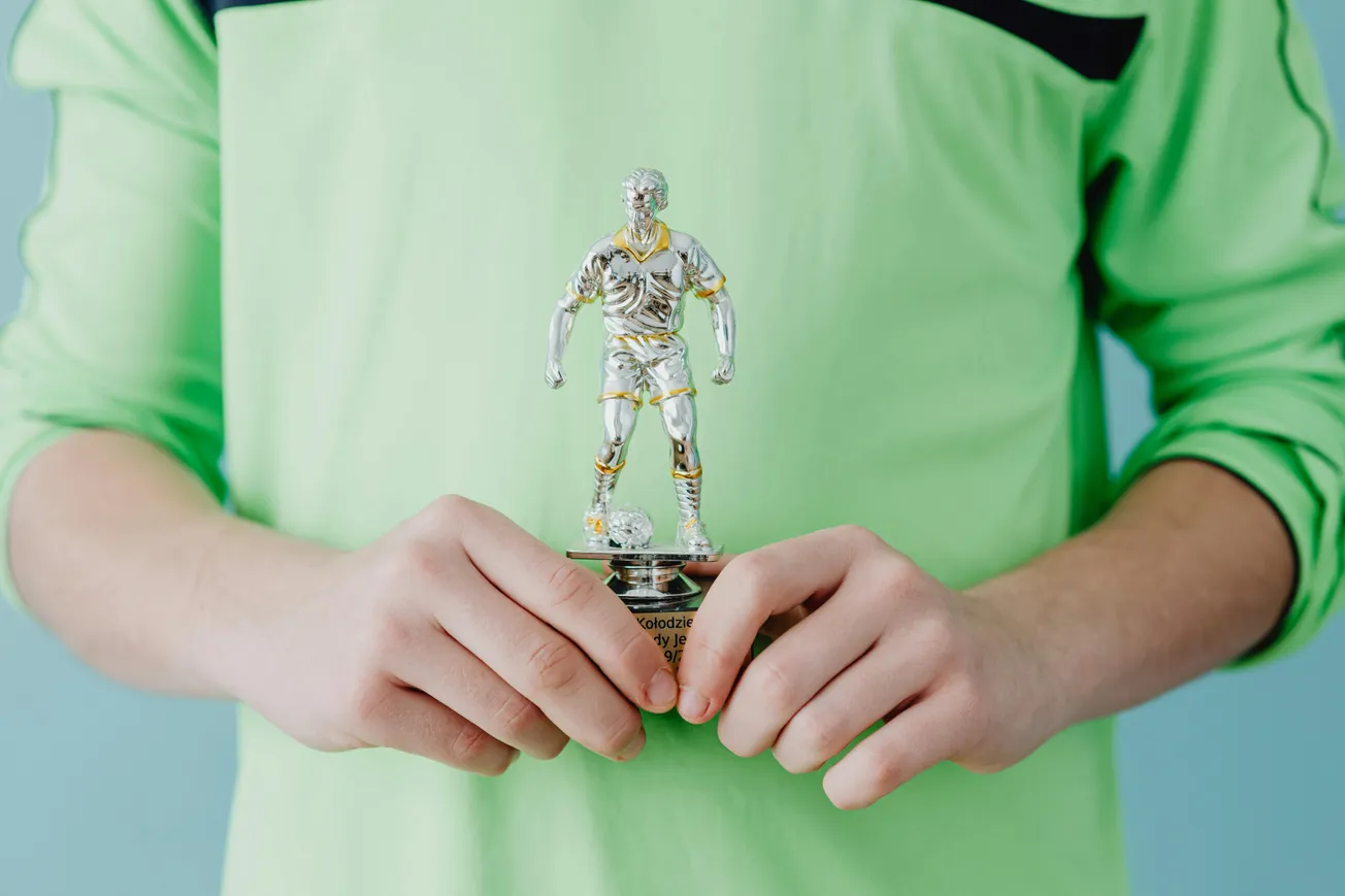 A person in a green jersey holds a small silver soccer trophy featuring a player, set against a light blue background. The mood is celebratory.