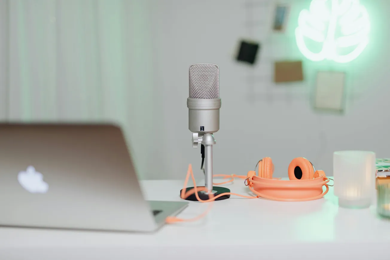 A podcast setup features a silver microphone, orange headphones, and a laptop on a white desk. A green leaf neon sign glows in the blurred background.