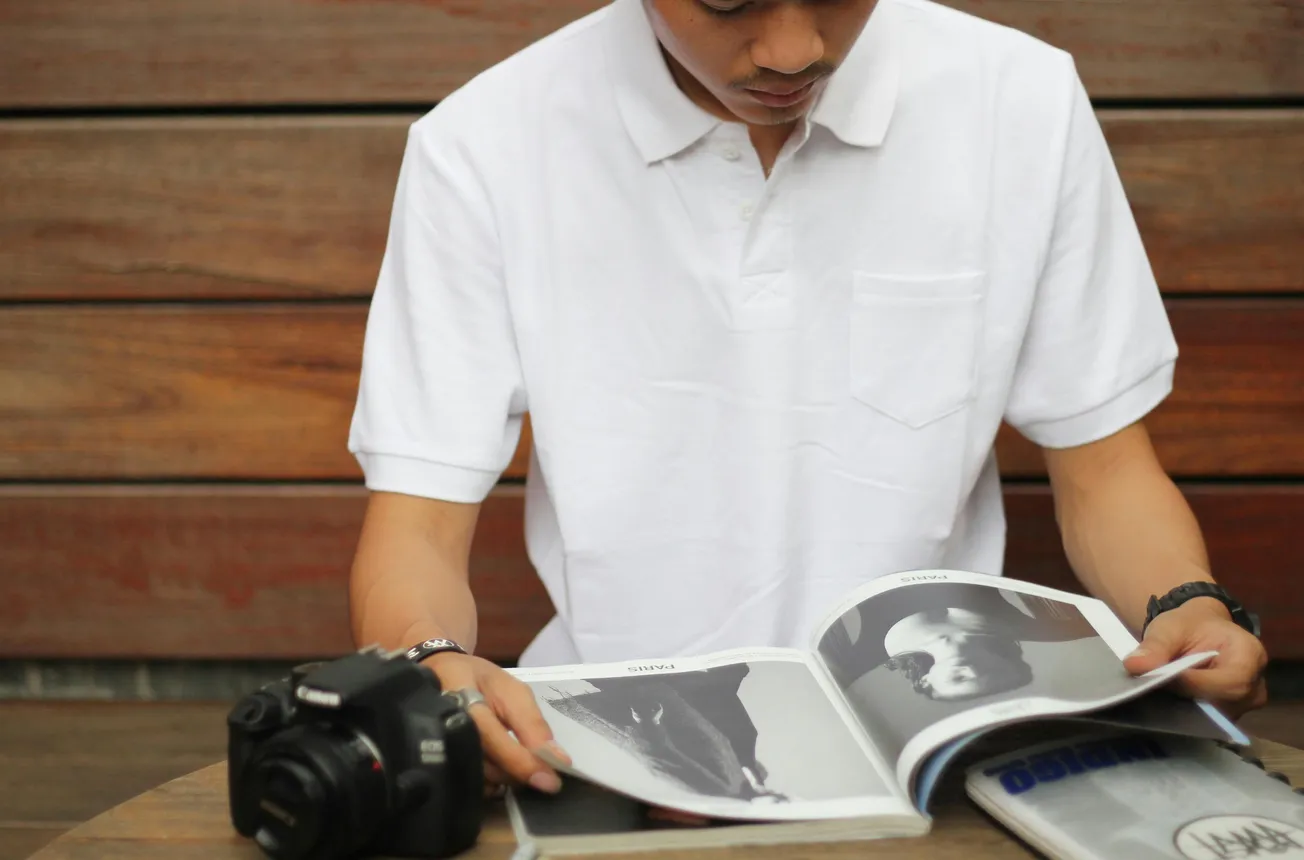 A person in a white polo shirt sits at a wooden table, intently reading a photography magazine. A camera rests beside them, conveying a calm, focused mood.