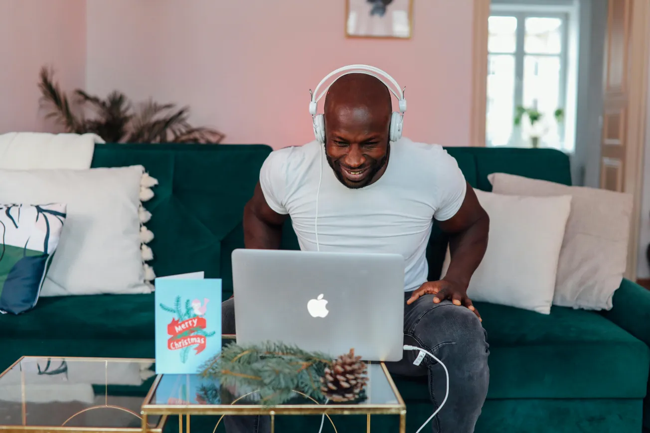 Man in a white t-shirt and headphones smiles at a laptop on a glass table with a "Merry Christmas" card and pinecones, sitting on a green sofa.