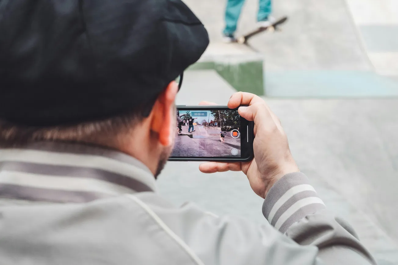A person in a cap films a skateboarder performing a trick with their phone at a skate park, with focused attention and dynamic action captured.