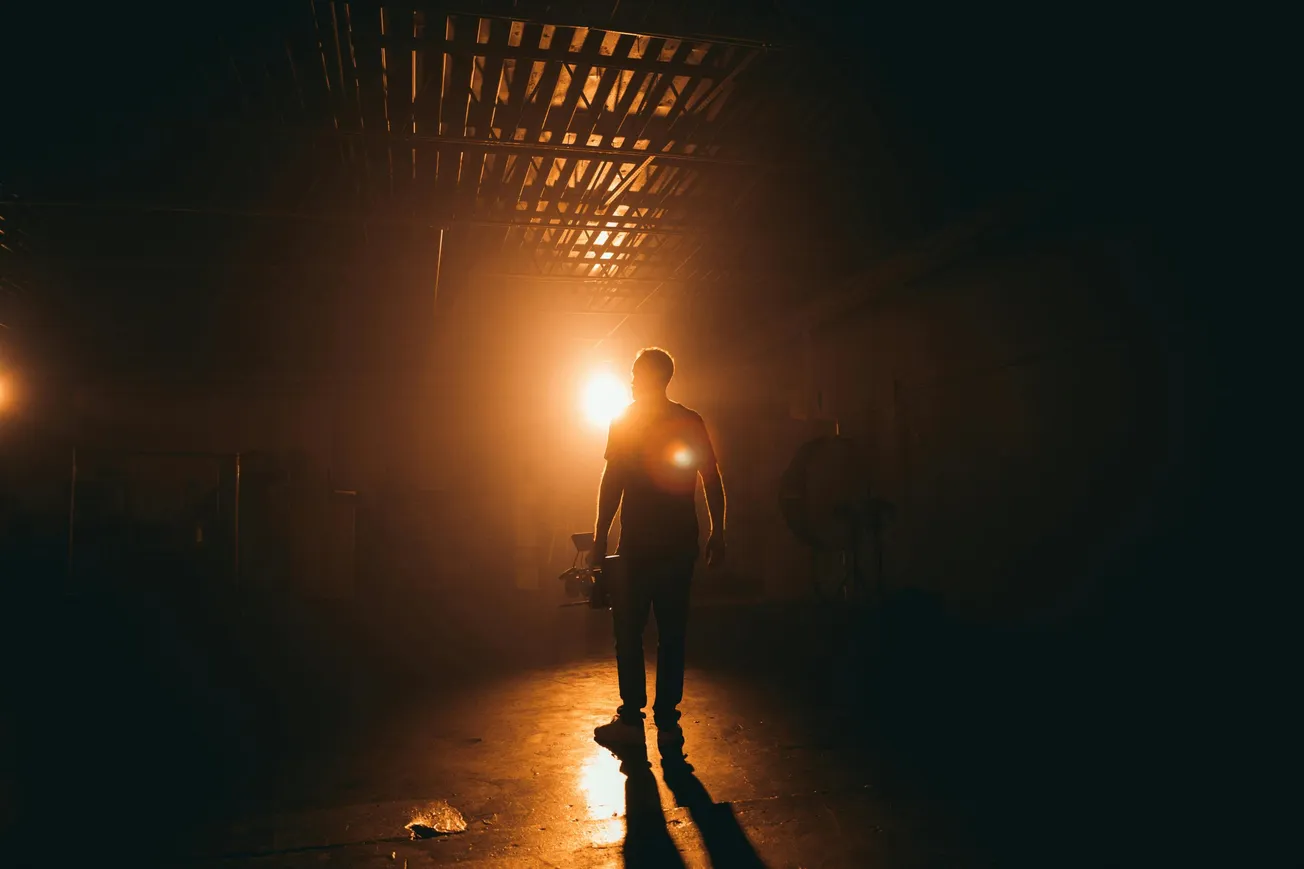Silhouetted person holding equipment stands in dimly lit room with ceiling grids. Two bright lights create a dramatic, mysterious atmosphere.