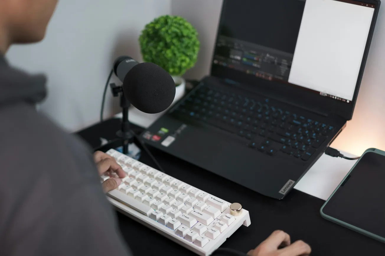 A person types on a white mechanical keyboard in front of a laptop, with a microphone nearby. A small plant adds a touch of green to the workspace.