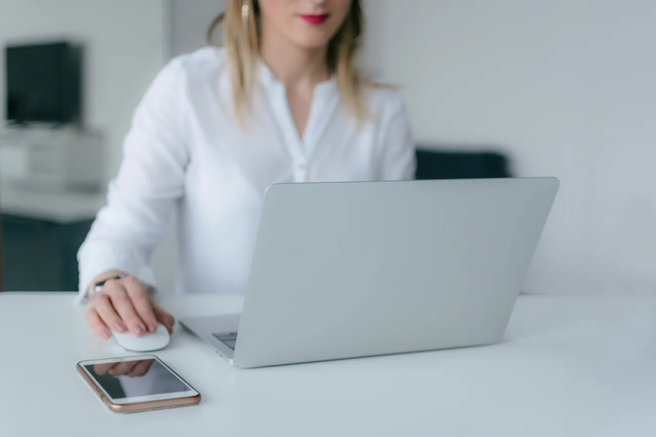 A woman in a white shirt uses a laptop on a white desk, with a smartphone nearby. The setting is a modern, minimalistic office, conveying focus.