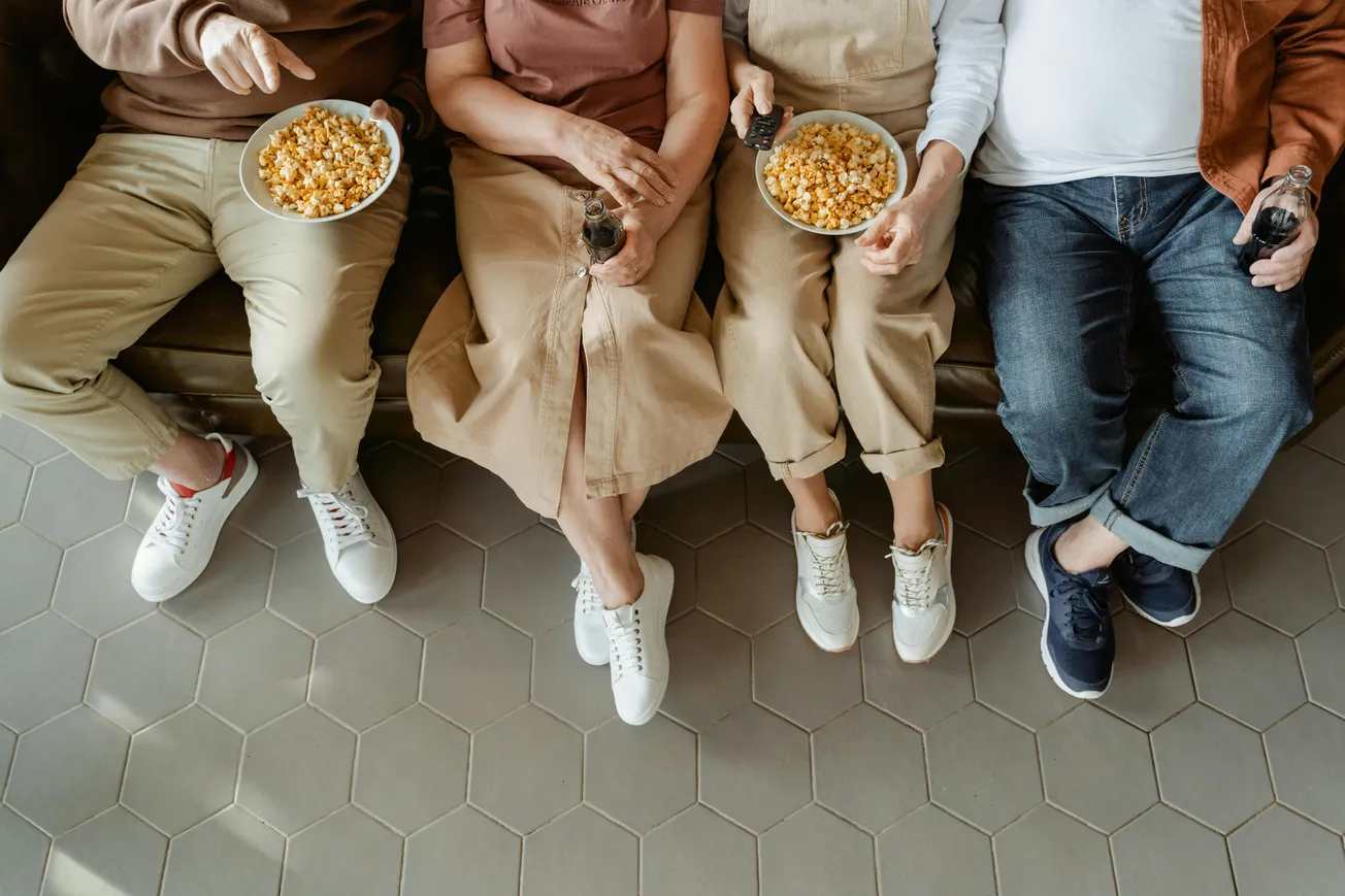 Four people sit on a couch, holding popcorn and drinks. They wear casual clothes and sneakers, conveying a relaxed, cozy atmosphere.