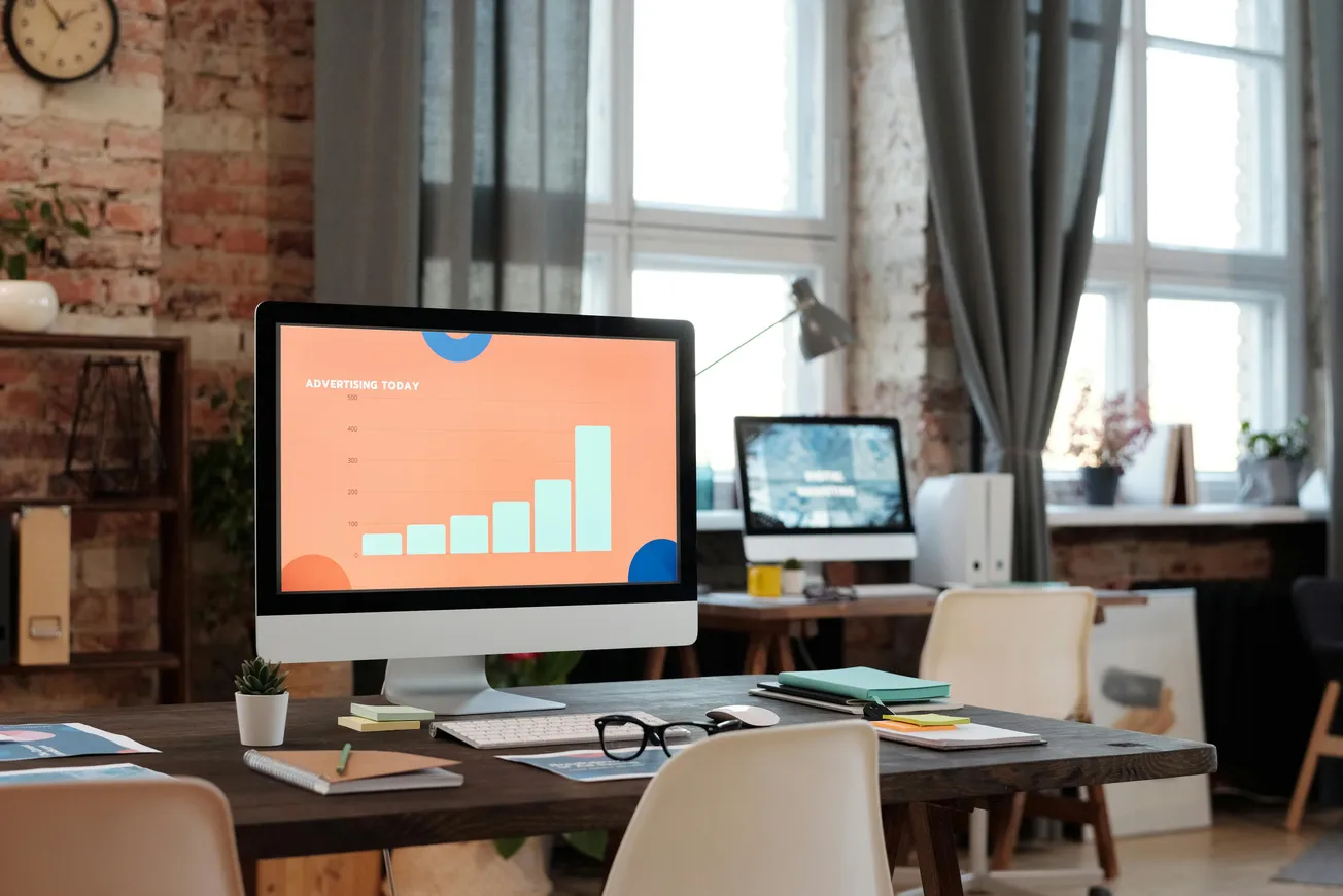 Modern office with a computer displaying a bar chart titled "Advertising Today" on a desk. Sunlight filters through large windows, creating a bright atmosphere.