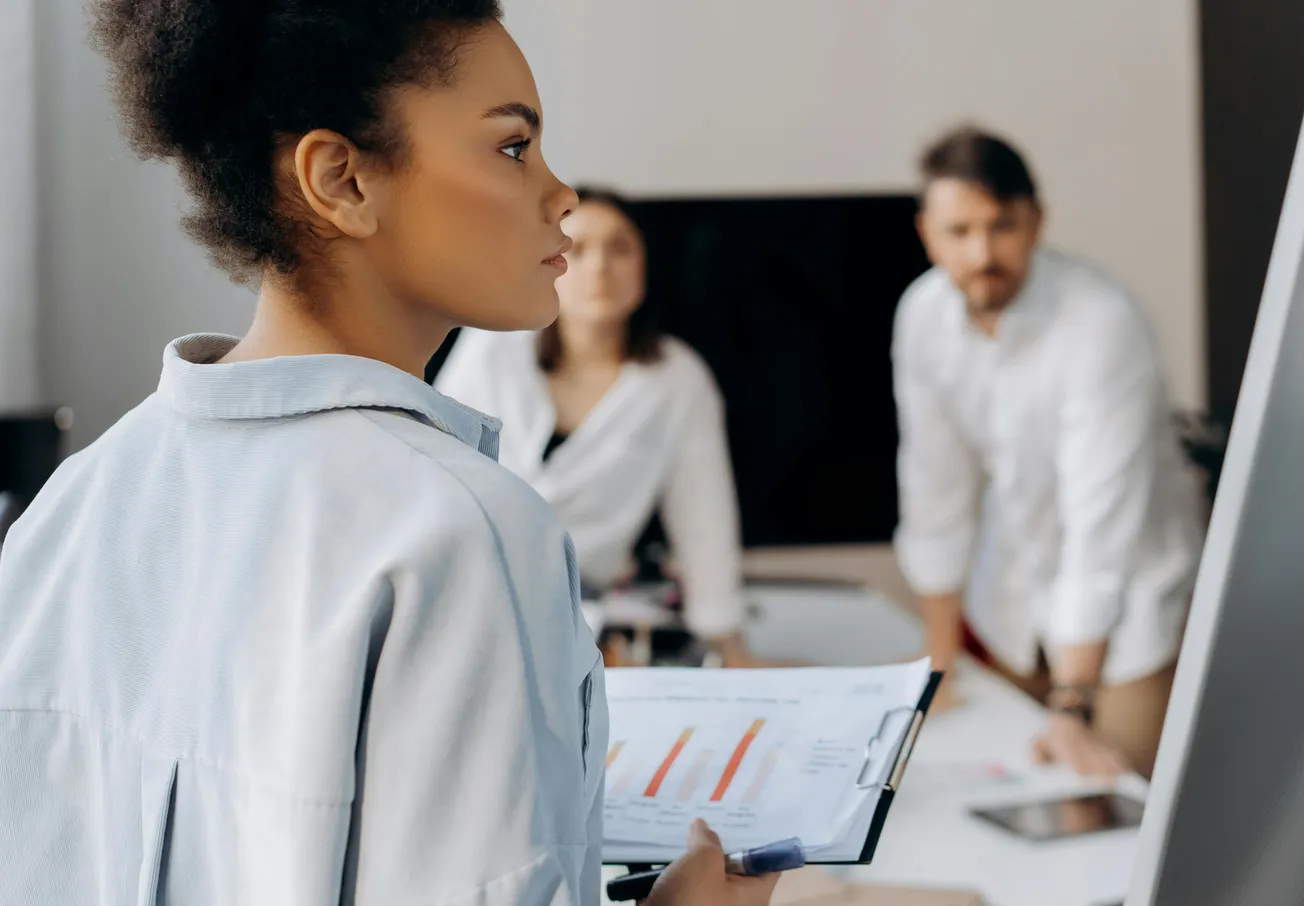 A woman presents data on a clipboard with a focused expression. Two colleagues in the background, both attentive, suggesting a professional meeting.