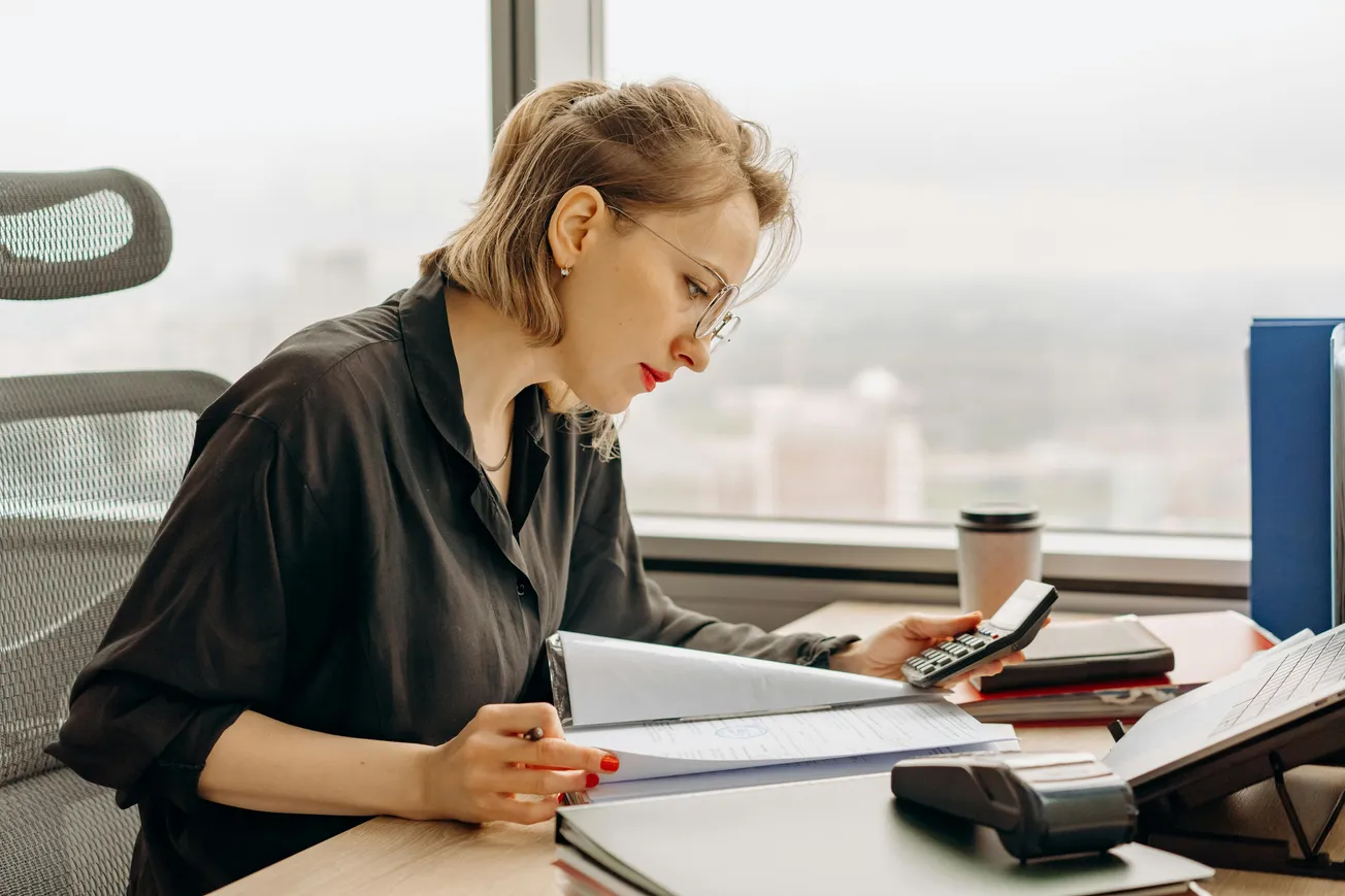 A woman in a black shirt is focused on work at a desk, using a calculator with one hand and reviewing a document in the other. A window view and coffee cup add to the office setting.