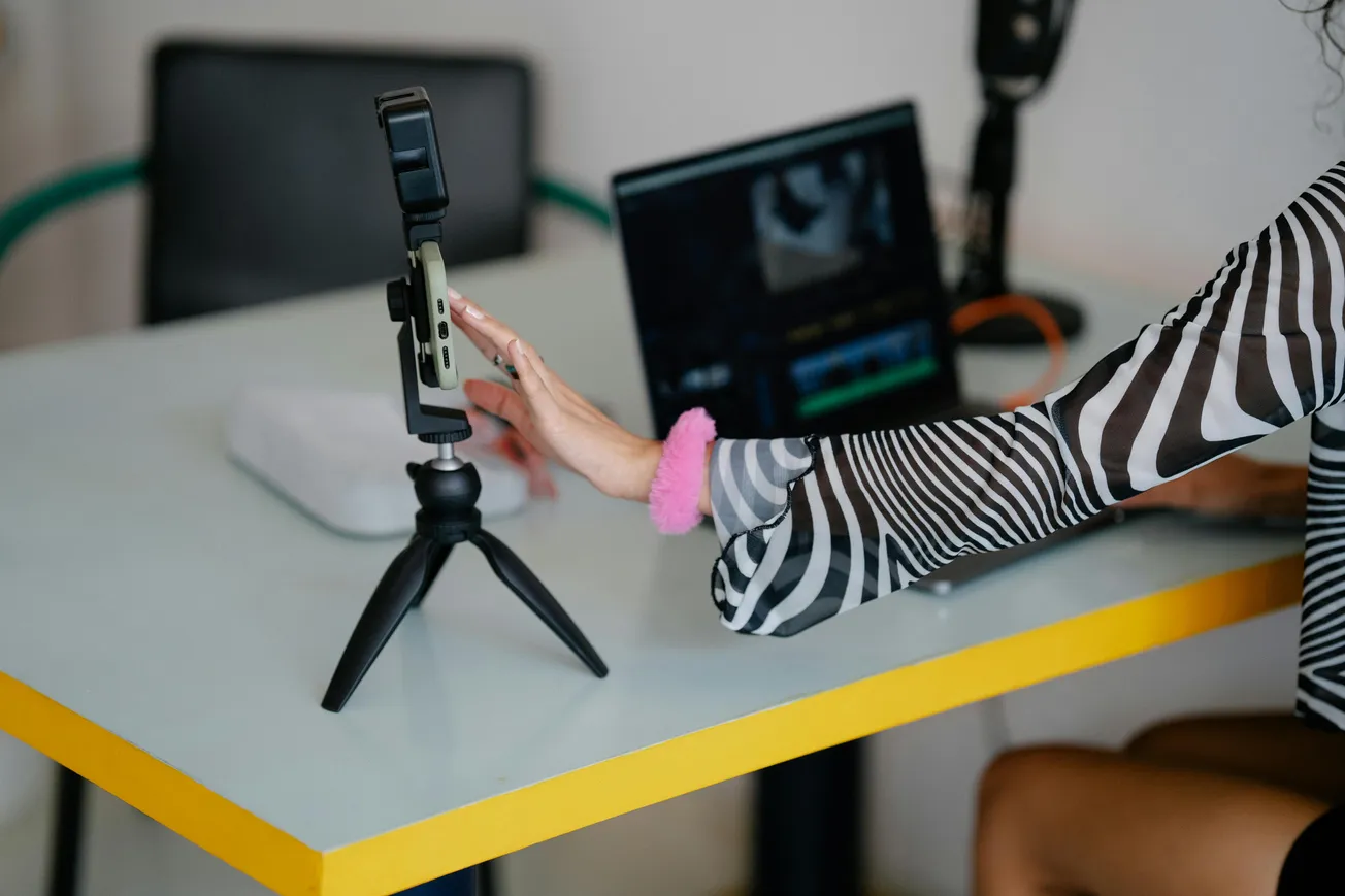 A person with a striped sleeve adjusts a smartphone on a tripod on a desk. Nearby are a laptop and microphone, suggesting a content creation setup.