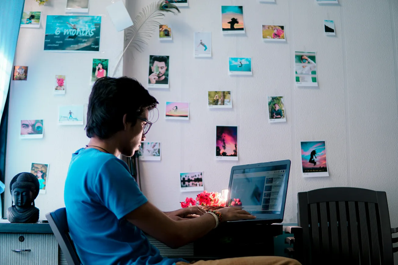 A person in a blue shirt types on a laptop in a cozy room. A wall behind is adorned with various colorful photos, exuding a creative, serene vibe.