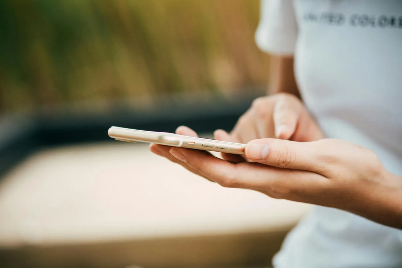 A person in a white shirt is using a smartphone outdoors. Their fingers are tapping the screen, conveying focus and engagement. Background is blurred.