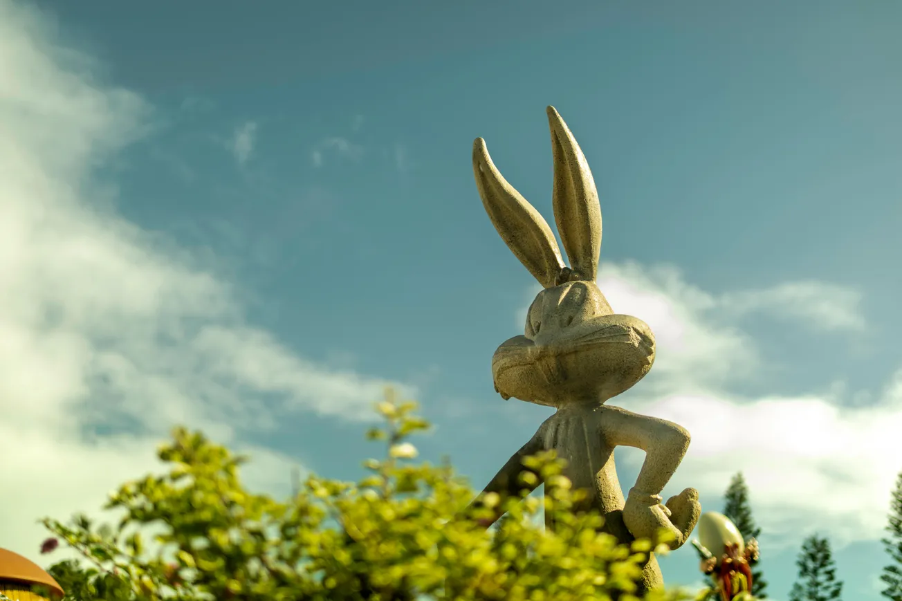 Statue of a cartoon rabbit stands amidst vibrant greenery under a clear sky. The rabbit is upright with large ears, adding a playful, whimsical feel.