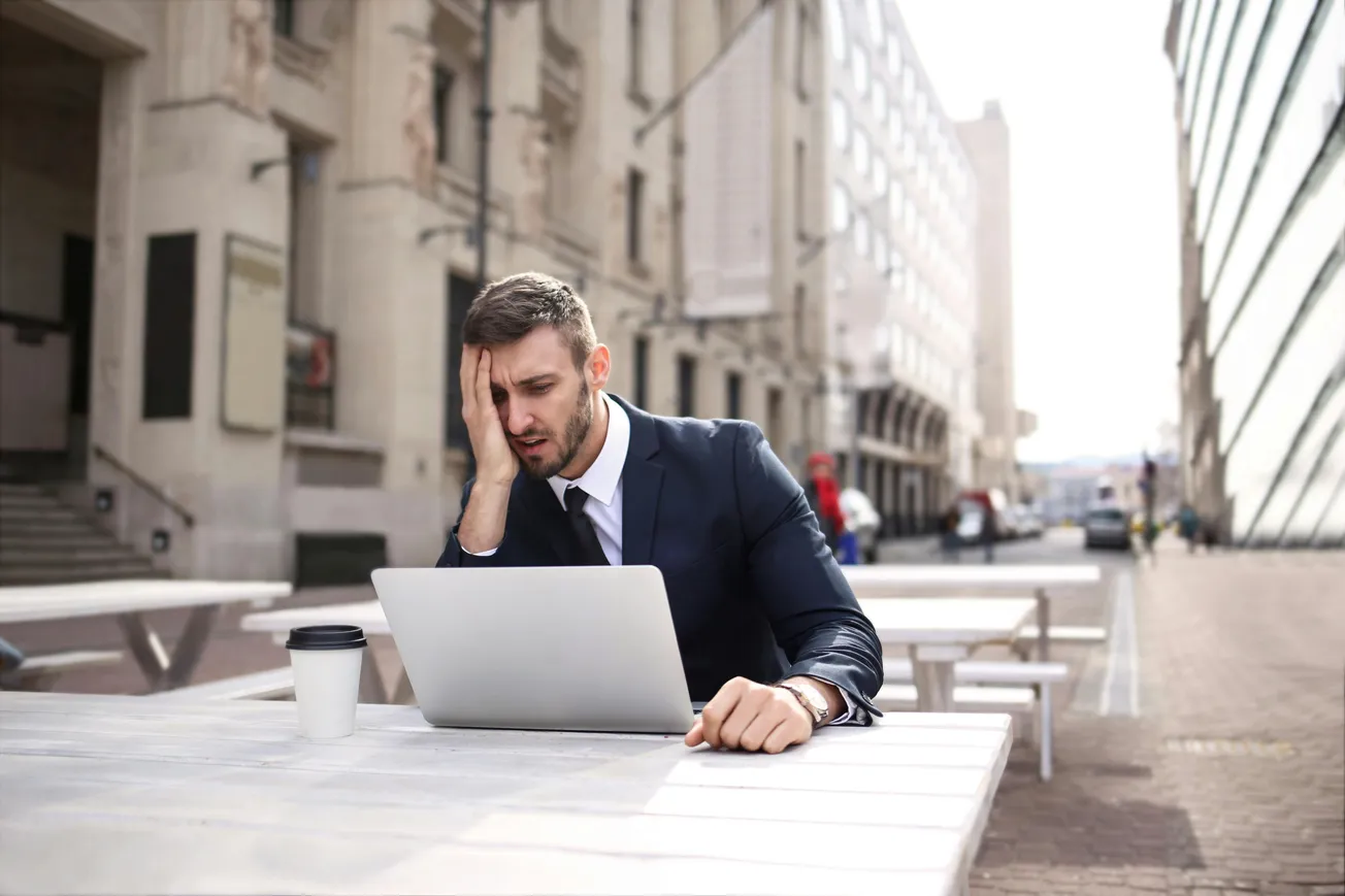 A man in a suit sitting at an outdoor table looks stressed, with one hand on his head, in front of a laptop. Coffee cup nearby. Urban setting.