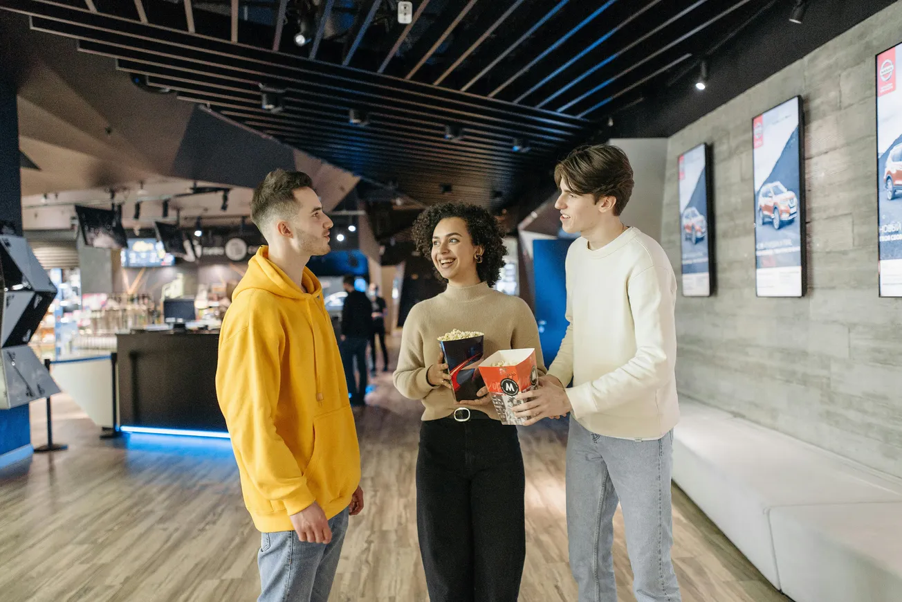 Three friends stand in a modern cinema lobby, chatting happily. One holds popcorn and drinks. The atmosphere is lively and inviting.
