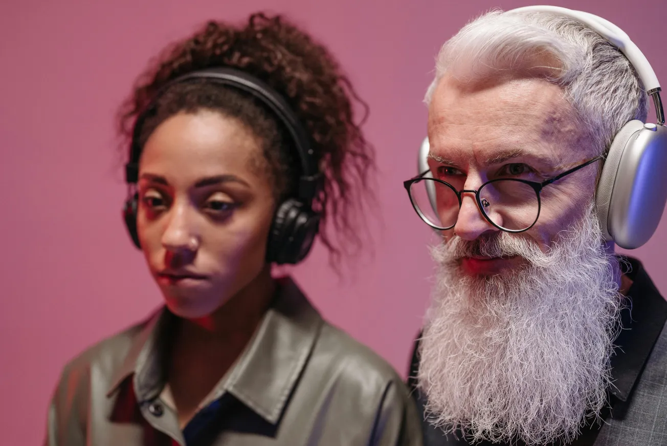 A woman with curly hair and a man with a long white beard, both wearing headphones, stand against a pink background, conveying a focused and calm mood.