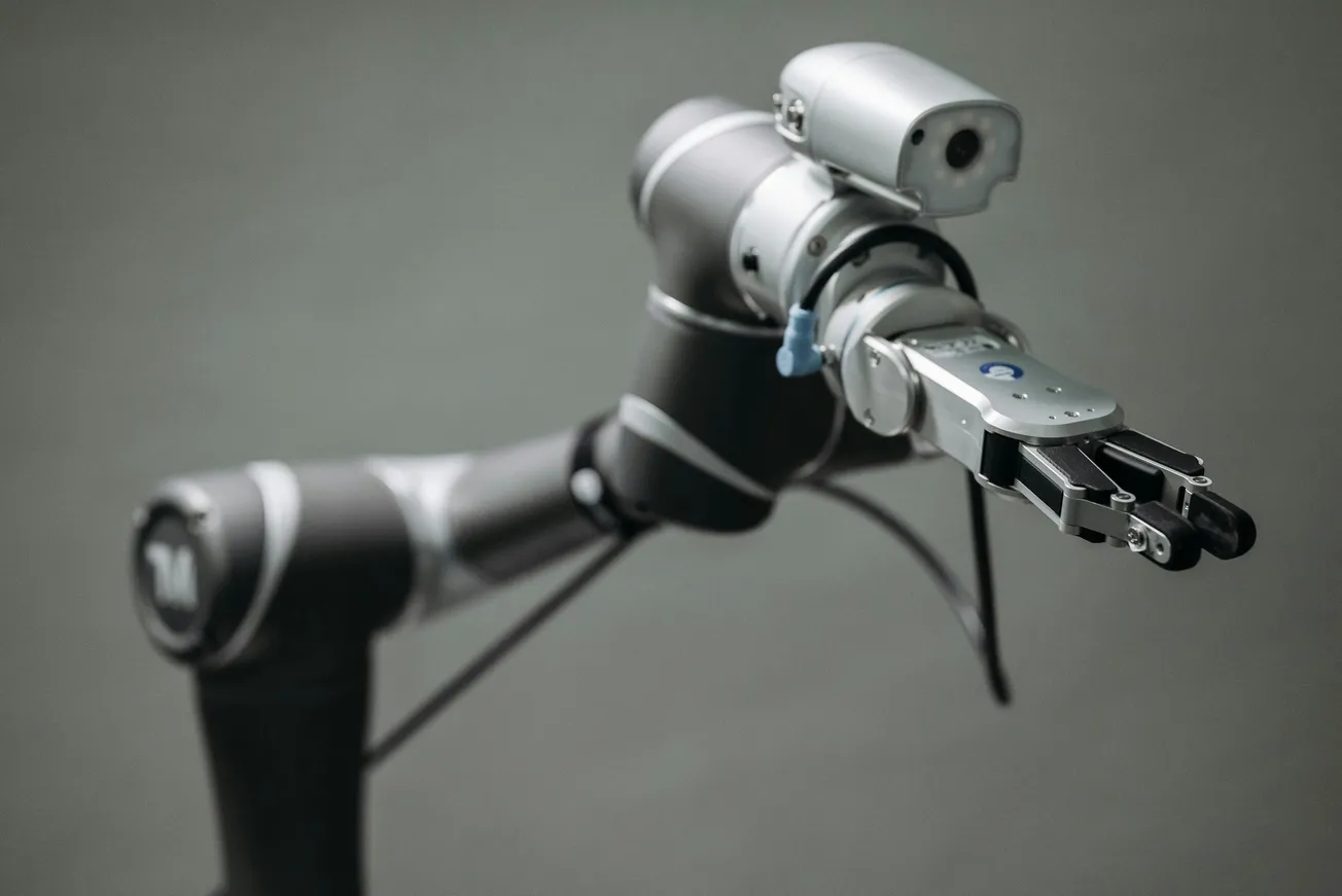 A sleek robotic arm extends forward, showcasing metallic joints and a precise gripper, set against a neutral gray background, conveying technological advancement.