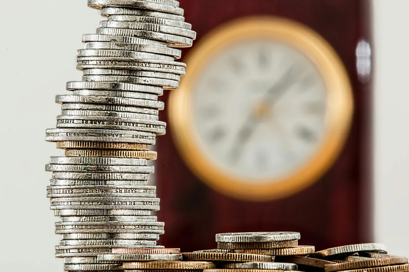 Stack of silver coins leaning to the left, with a blurred, round clock in the background. Represents time and money relationship, conveying a sense of imbalance.