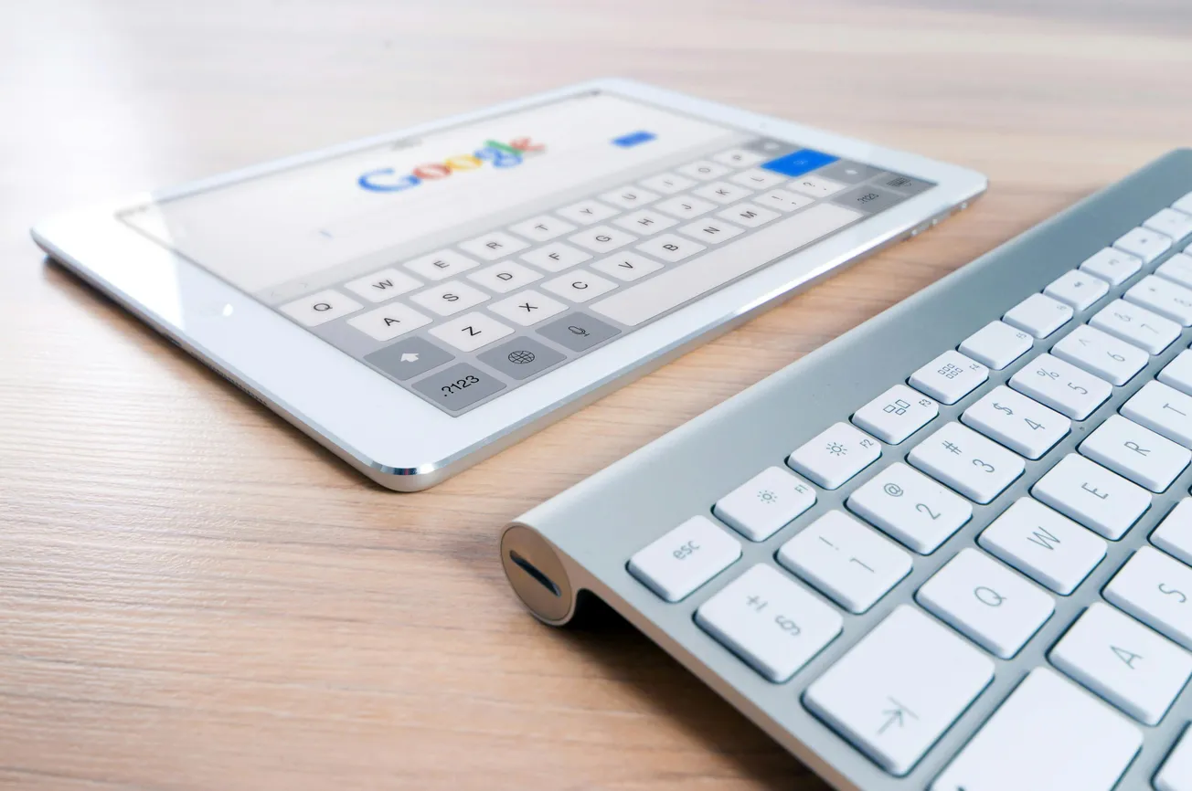 A silver keyboard and a tablet displaying the Google search page rest on a wooden surface, conveying a tone of digital connectivity and modern technology.