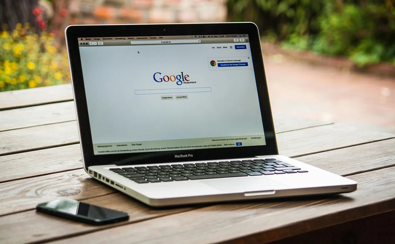 A MacBook Pro displaying the Google homepage is on a wooden table outdoors. A smartphone lies beside it. The setting feels relaxed and natural.