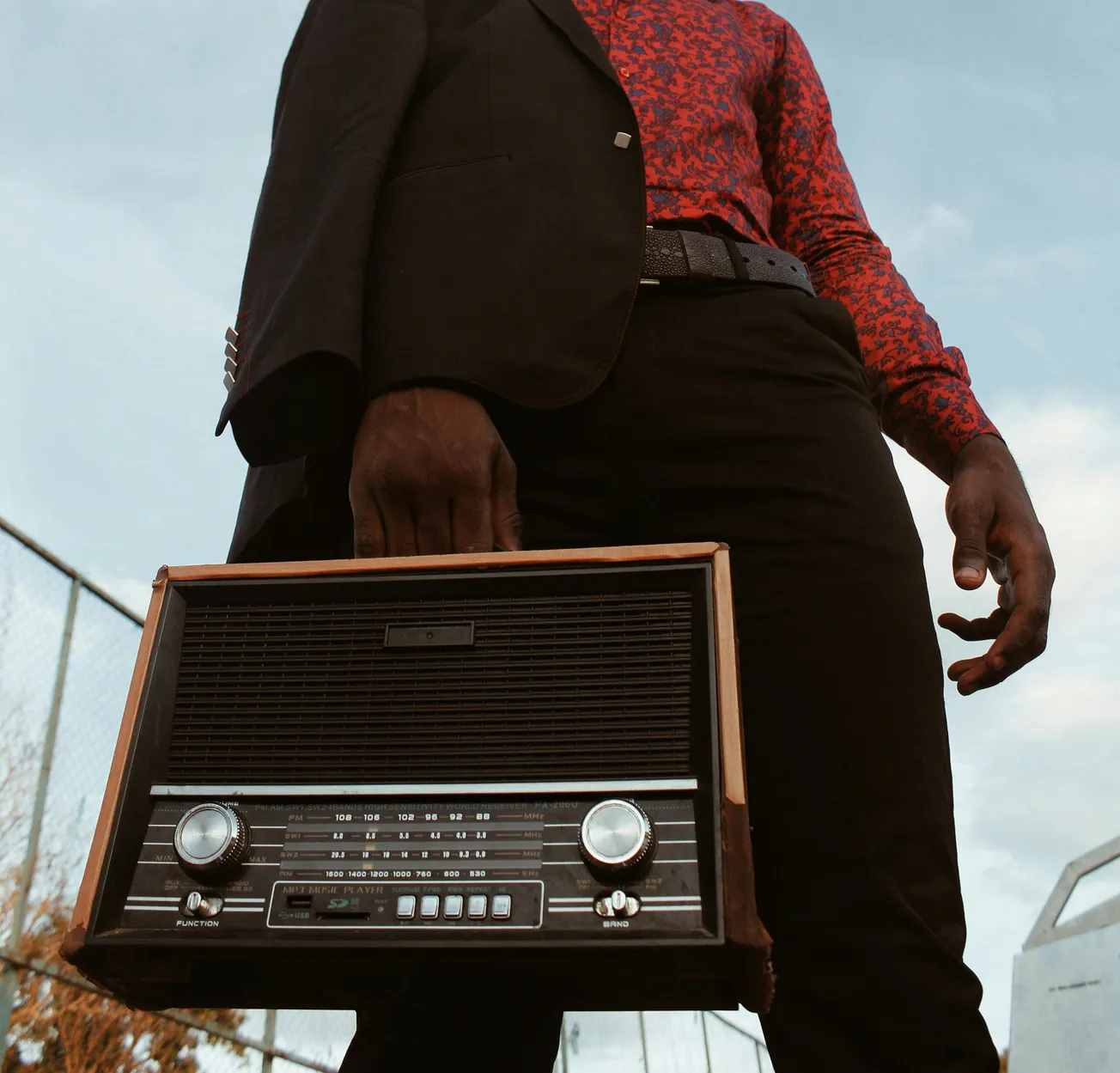 Man in a red patterned shirt and black suit holds a vintage radio against a blue sky backdrop. The image conveys a retro and stylish vibe.