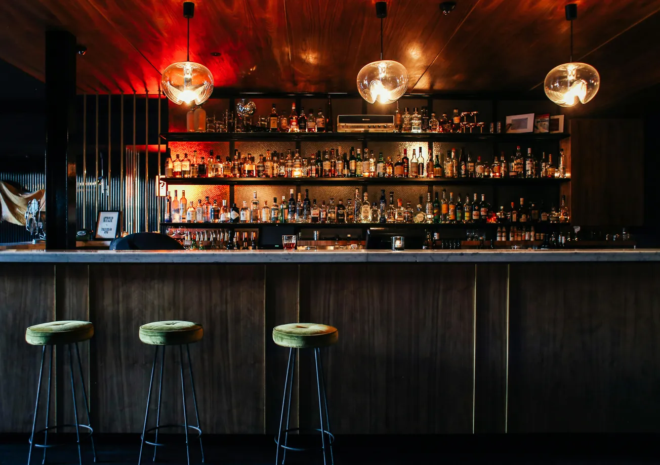 Warmly lit bar scene with wooden shelves stocked with bottles, globular pendant lights, and three green-cushioned stools. The ambiance is cozy and inviting.