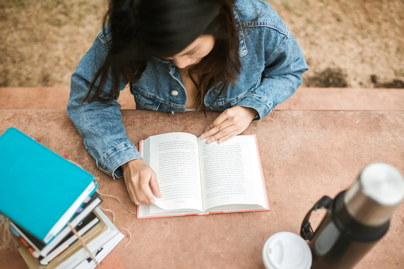 A person in a denim jacket reads an open book on a stone table outdoors. Nearby, a stack of books and a thermos with a cup denote a study scene.