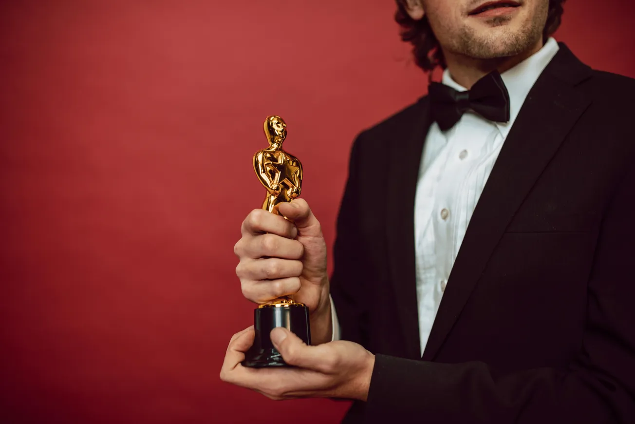 Man in a tuxedo holding a gold award statuette against a red background, conveying a sense of achievement and celebration.