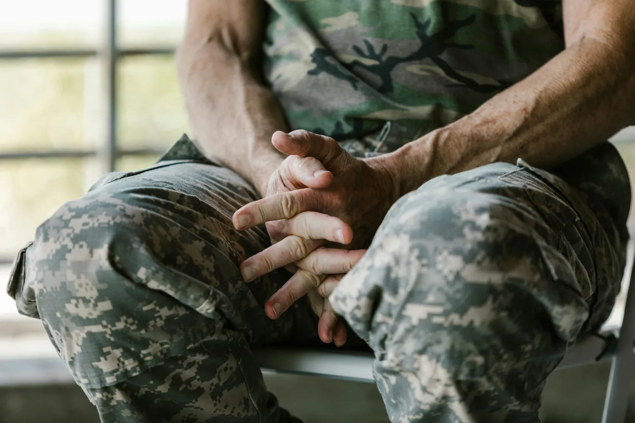 A person in camouflage clothes sits with fingers interlocked, creating a contemplative tone. Sunlight filters through a window, highlighting muscular arms.
