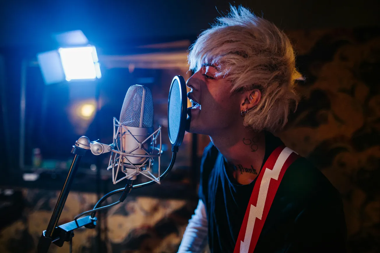 Musician with platinum blond hair passionately sings into a studio microphone. Warm, moody lighting creates an intense and creative atmosphere.