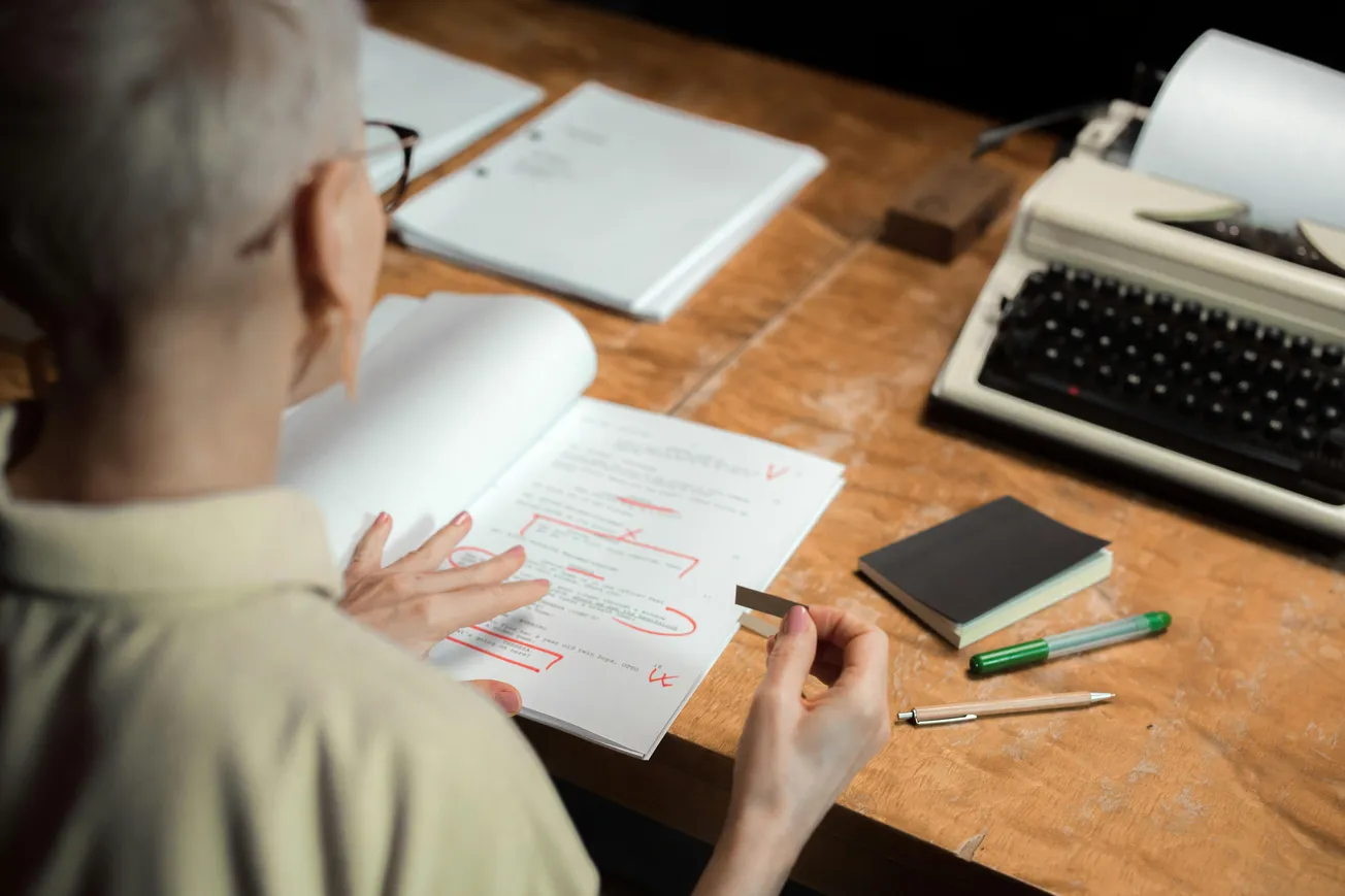 A person with short hair reviews a marked-up document with red edits. Nearby lie a typewriter, closed notebook, and pens on a wooden desk. The scene conveys focus.