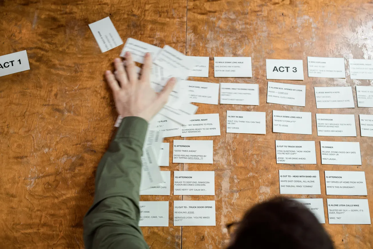 A person organizes note cards on a wooden table labeled "Act 1" and "Act 3," indicating a project or script planning session. Focus and creativity are conveyed.