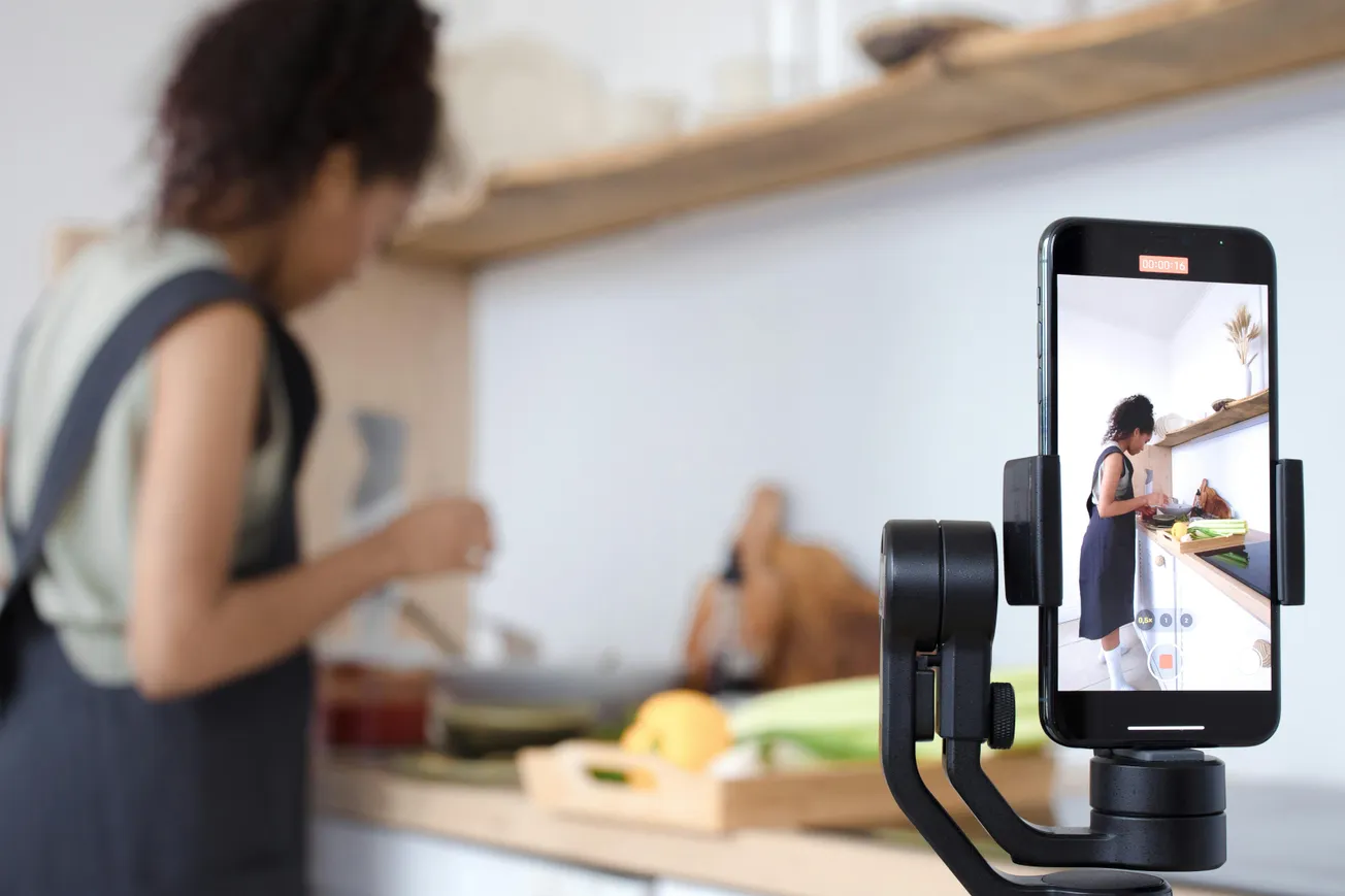 A woman in a blurred background prepares food in a kitchen. In the foreground, a smartphone on a tripod records her, showing her chopping vegetables.
