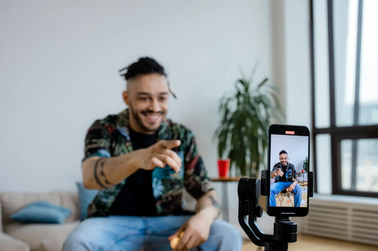 A smiling man sits on a couch, joyfully gesturing towards a smartphone on a tripod, capturing a video. A red cup and plant are in the background.