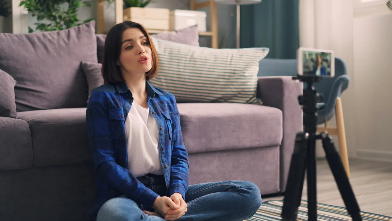 A woman in a blue shirt sits cross-legged on the floor, recording herself with a smartphone on a tripod. She appears relaxed, couch in the background.