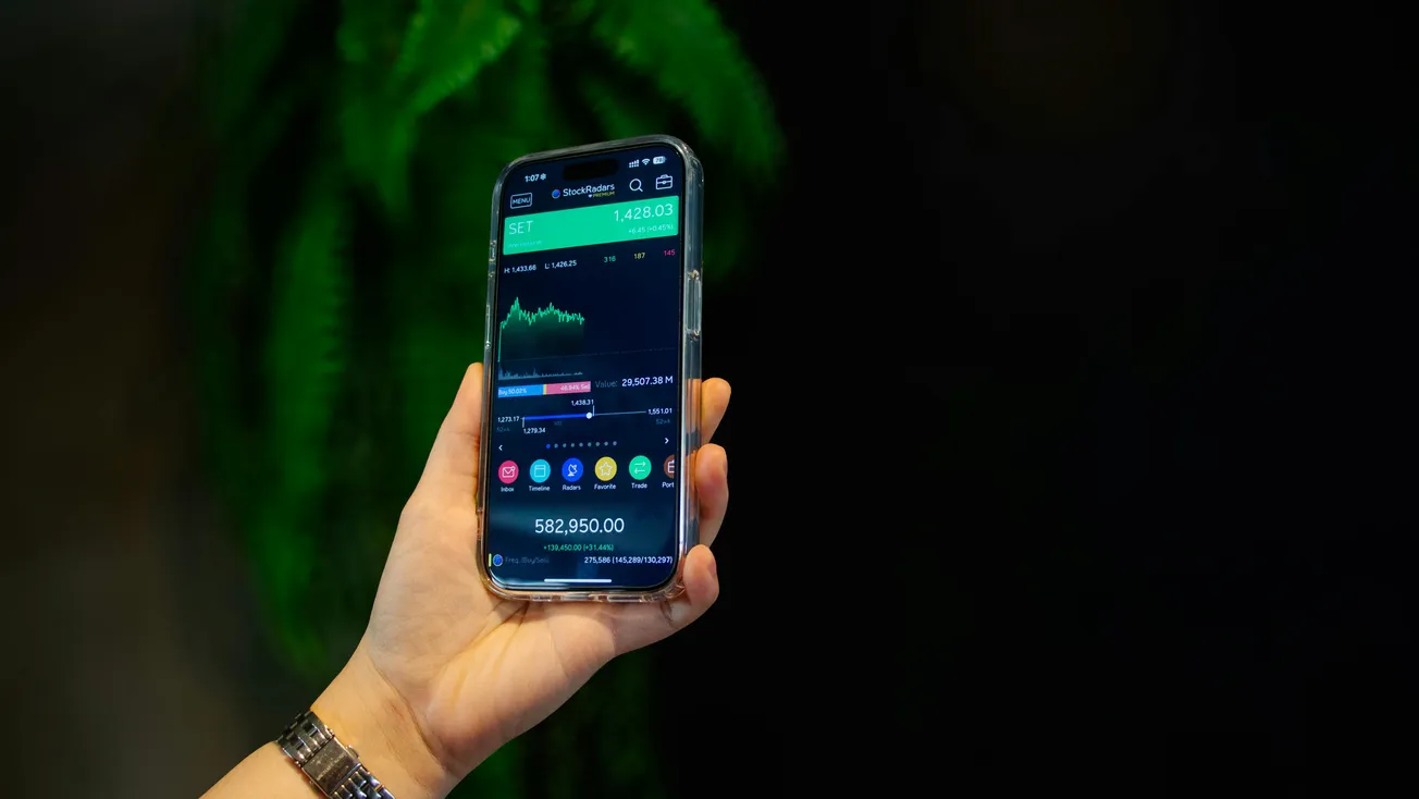 A hand holds a smartphone displaying a stock market app with graphs and numbers. The background is dark with green leaves, suggesting a focused, analytical mood.