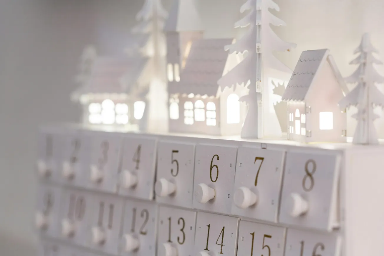 A white Advent calendar with numbered drawers and small illuminated houses and trees on top, evoking a serene, festive winter village scene.