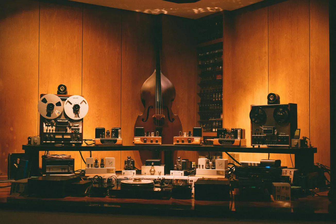 A cozy room with a vintage audio setup on a wooden shelf, featuring reel-to-reel tape decks and an upright bass in the background. Warm lighting creates a nostalgic feel.