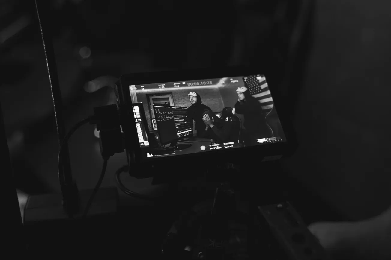 Black and white shot of a video camera screen showing three people in a room, possibly filming. An American flag is visible in the background.